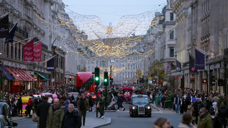 Crowds-of-shoppers-fill-the-pavements-on-Regent-Street-as-black-Hackney-taxi-cabs,-red-buses-and-brightly-lit-rickshaw-cabs-under-the-festive-lights-at-dusk