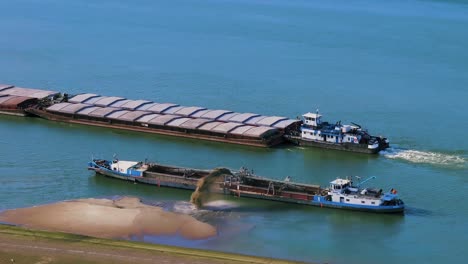 Aerial-close-up-shot-of-a-dredger-unloading-dredged-sand-on-a-big-river,-other-ships-passing-by,-sunny-day