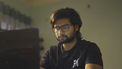 A-South-Asian-Indian-adult-with-beard-wearing-glasses-typing-or-working-on-a-laptop-with-a-serious-face-in-a-warm-environment-room