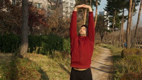 Mid-Adult-Man-Active-Exercising-Outdoors-in-a-Park-Trail-Stretching-Torso-Locking-Arm-Above-Head-and-Jumping-with-Warming-up-Shoulders-Rotations-Outdoors-at-Apartment-Park-in-Autumn---slow-motion