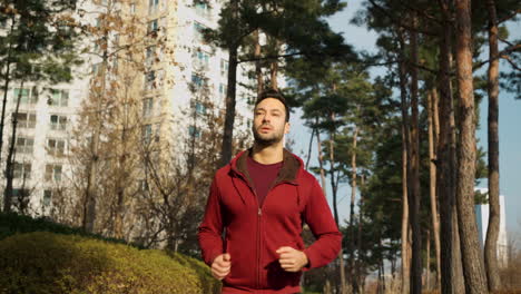 Attractive-man,-guy-jogging-in-the-park-on-a-background-of-pine-trees-and-modern-apartments---low-angle
