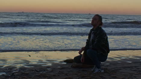 caucasian-male-man-model-sits-squats-on-the-beach-near-the-sea-ocean-on-the-sand-shore-with-sunset-and-cinematic-actor's-play