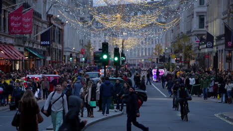 People-with-shopping-bags-cross-the-street-between-black-Hackney-taxi-cabs,-bicycles-and-brightly-lit-rickshaw-cabs-under-the-festive-lights-on-Regent-Street-at-dusk