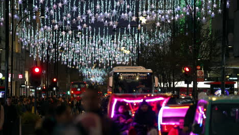 In-Zeitlupe-Radeln-Rikscha-Taxis-Mit-Hellen-Lichtern-Durch-Den-Verkehr,-Während-Weihnachtseinkäufer-Nachts-Die-Bürgersteige-Unter-Den-Festlichen-Lichtern-Der-Oxford-Street-Füllen