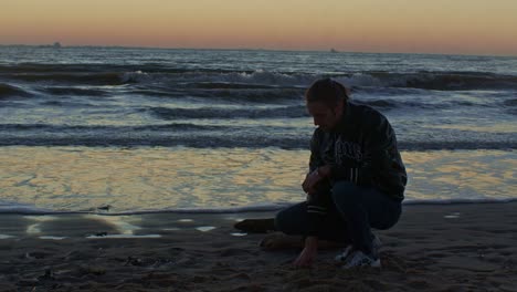 caucasian-male-man-model-sits-squats-and-grabs-the-sand-with-his-hand-on-the-beach-near-the-sea-ocean-on-the-sand-shore-with-sunset-and-cinematic-actor's-play
