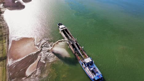 Aerial-top-shot-of-a-dredger-unloading-dredged-sand-on-a-big-river,-sunny-day