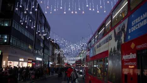 Eine-Reihe-Roter-Busse-Im-Verkehr-Fährt-Los,-Während-Weihnachtseinkäufer-Nachts-Die-Bürgersteige-Unter-Den-Festlichen-Lichtern-Der-Oxford-Street-Füllen