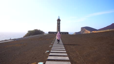 Woman-in-pink-jacket-walks-towards-Lighthouse-of-Ponta-dos-Capelinhos-surrounded-by-volcanic-ashes,-rear-shot