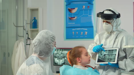 Dentist-in-protective-equipment-showing-on-tablet-dental-x-ray-of-child
