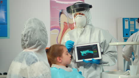 Dentist-with-ppe-suit-pointing-on-digital-screen-explaining-x-ray-to-mother