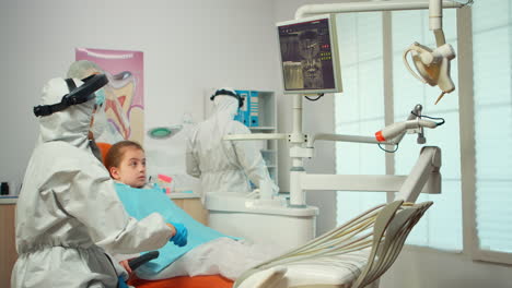 Dentist-in-ppe-suit-pointing-on-digital-dental-monitor-explaining-x-ray-to-mother