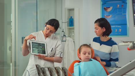 Odontólogo-Pediátrico-Mostrando-Rayos-X-De-Dientes-En-La-Pantalla-De-Una-Tableta-A-La-Madre