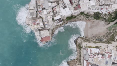 Top-down-aerial-footage-of-the-waves-washing-up-against-the-rocks-and-shore-in-Polignano-a-Mare,-Italy-on-a-stormy-day