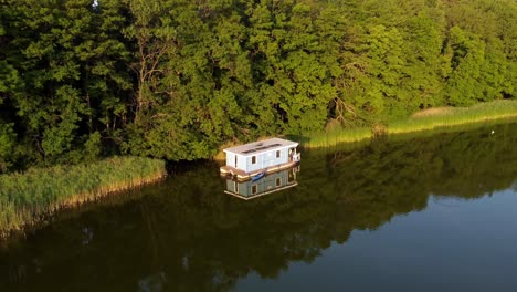 Houseboat-floating-on-a-lake-next-to-a-forest-during-sunset-in-Brandenburg,-Germany