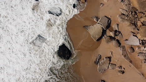 Aerial-top-down-shot-of-foamy-waves-reaching-golden-beach-with-Rocks-in-Portugal-at-sunny-day