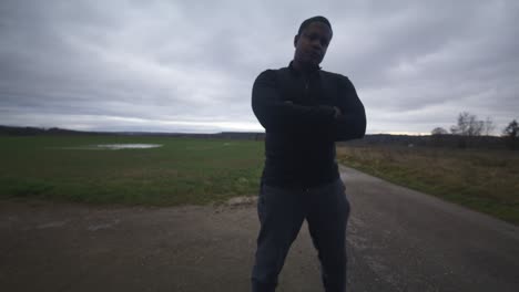 Modern-Black-Male-Waiting-in-Open-Field-Under-Grey-Sky,-Europe,-4K-|-Muscular,-Intimidating,-Shadow,-Figure,-Blurry,-Dark-Clothes,-Powerful,-Shrug,-Bully,-Blocking,-Obstacle,-Mean,-Angry,-Arms-Crossed