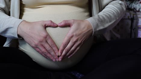 Mujer-Embarazada-Se-Toma-De-La-Mano-En-Forma-De-Corazón-Cerca-De-Luces-Navideñas-Centelleantes