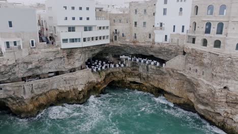 Aerial-footage-of-a-restaurant-in-Polignano-a-Mare-that-is-located-inside-a-cave-in-the-cliffs-with-the-waves-of-the-Mediterranean-Sea-crashing-below