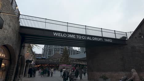 Looking-Up-At-Signage-Saying-Welcome-To-Coal-Drops-Yard-With-Shoppers-Walking-Past-On-Overcast-December-Day