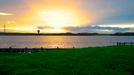 Zeitraffer-Eines-Wunderschönen-Sonnenuntergangs-über-Einem-See-Mit-Ruhiger-Wasseroberfläche-Und-Einer-Windkraftanlage-In-Der-Ferne