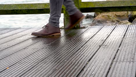 Close-up-view-of-man's-legs-in-jeans-and-brown-leather-boots-walking-on-a-wooden-pier
