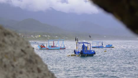 Asian-fishing-boats-bobbing-on-gentle-ocean-waves-with-mountains-in-backgrounds-and-rocks-in-foreground,-filmed-in-handheld-style-as-medium-shot