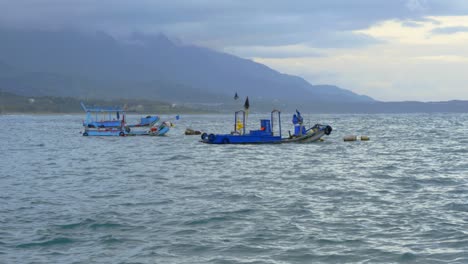Anchored-asian-fishing-vessels-bobbing-in-gentle-ocean-swell-with-mountains-and-cloudy-sky-in-background,-filmed-as-stationary-medium-wide-shot