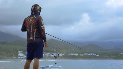Hoodie-wearing,-cigarette-smoking-fisherman-holding-a-fishing-rod-and-reel-with-mountains-and-gray-clouds-in-background,-filmed-as-stationary-medium-wide-shot