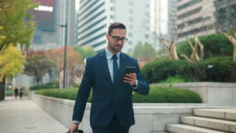 Confident-adult-bearded-businessman-in-suit-walking-near-modern-business-office-building-holding-smartphone-in-hands