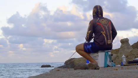 Man-wearing-hoodie-with-fishing-rod-pointed-towards-water-with-ocean-and-clouds-in-background,-filmed-from-backside-as-stationary-medium-shot