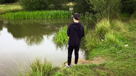 A-young-boy-stood-by-himself-at-the-edge-of-the-lake-fishing-on-a-dull-day-during-summertime