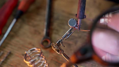 Close-up-of-the-hands-of-a-Caucasian-man-using-a-soldering-tin-iron-while-soldering-some-electrical-contacts-while-using-a-magnifying-glass