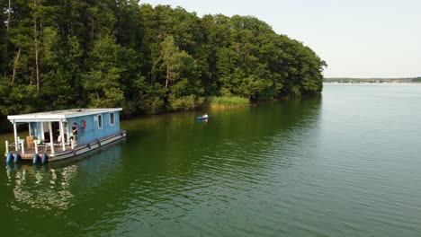 Person,-Die-Mit-Dem-Kanu-Zu-Einem-Hausboot-Auf-Einem-Grünen-Natursee-Neben-Einem-Wald-In-Brandenburg,-Deutschland,-Fährt