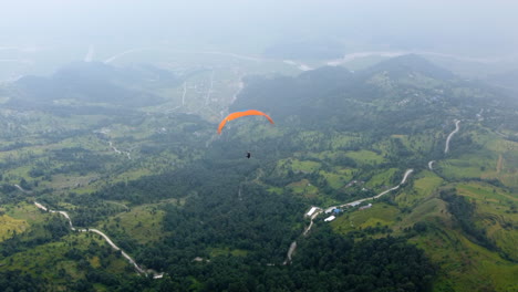 Paraglider-soars-with-orange-nylon-wing-above-beautiful-lush-green-valley-with-clouds-and-mist