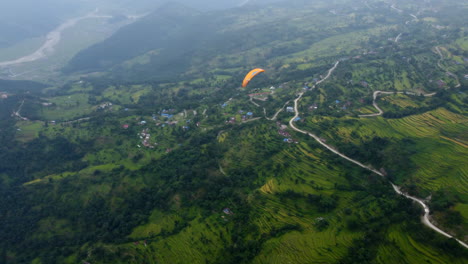 Paraglider-soars-and-drops-down-above-terraced-fields-as-raptor-bird-flies-above,-high-angle-aerial-overview