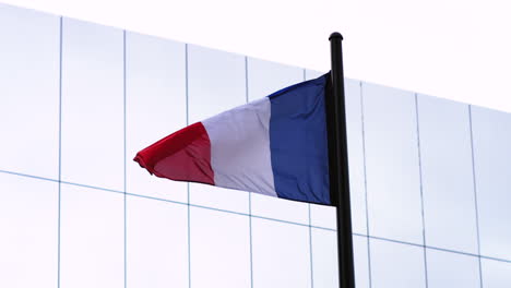 Zeitlupenaufnahmen-Der-Französischen-Flagge,-Die-Sich-Im-Wind-Bewegt,-Mit-Einem-Gespiegelten-Gebäude-Im-Hintergrund