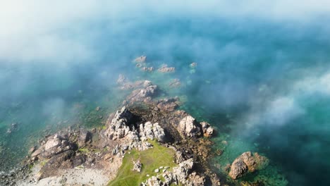 Mist-coming-in-off-the-sea-over-rocky-promontory-with-crystal-clear-blue-water-bright-overhead-shot-with-slow-moving-mist