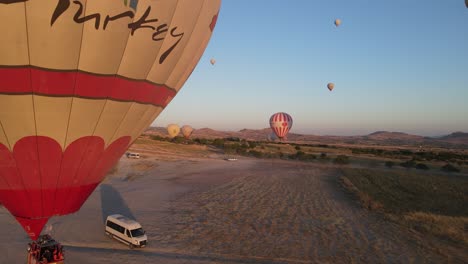 Drone-view-of-a-colorful-hot-air-balloon-inflated-on-a-wide-plain,-a-very-famous-tourist-attraction-in-Cappadocia,-Turkey,-the-moment-tourist-vehicles-approach-the-balloon