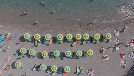 Top-down-aerial-footage-of-the-waves-of-the-sea-rolling-up-to-the-beach-with-tourists-and-beach-umbrellas