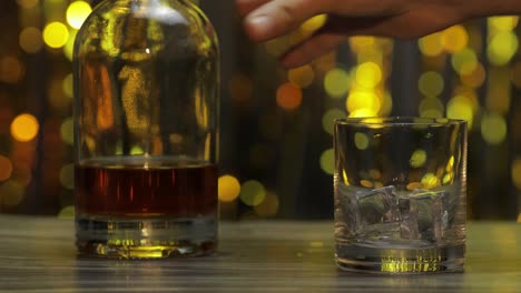 Gießen-Von-Goldenem-Whiskey,-Cognac-Oder-Brandy-Aus-Der-Flasche-In-Ein-Glas-Mit-Eiswürfeln.-Glänzender-Hintergrund