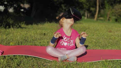 Child-in-VR-headset-helmet-sitting-in-lotus-position-on-mat-and-performing-yoga-meditation-in-park