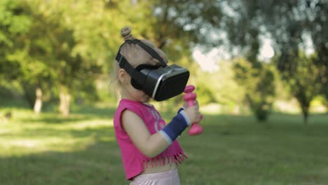 Athletic-child-girl-in-VR-headset-helmet-making-fitness-workout-exercises-with-dumbbells-in-park