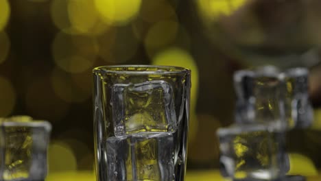 Barman-pour-frozen-vodka-from-bottle-into-shot-glass.-Ice-cubes-against-shiny-gold-party-background