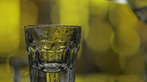 Barmann-Gießt-Gefrorenen-Wodka-Aus-Der-Flasche-In-Schnapsglas-Vor-Glänzendem-Goldparty-Hintergrund.-Nahaufnahme