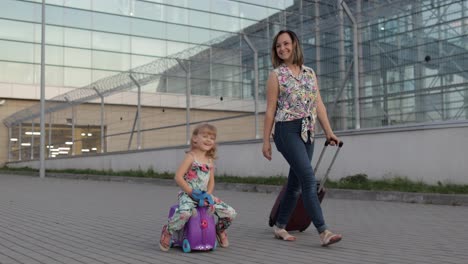 Mother,-daughter-walking-from-airport-after-vacation.-Woman-carrying-luggage.-Child-rides-suitcase