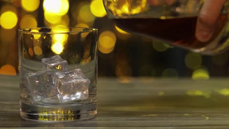 Pouring-of-golden-whiskey,-cognac-or-brandy-from-bottle-into-glass-with-ice-cubes.-Shiny-background
