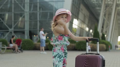 Child-girl-tourist-with-suitcase-bag-near-airport.-Kid-with-luggage-dances,-rejoices,-show-thumbs-up