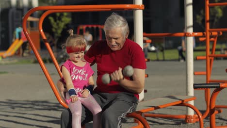 Granddaughter-and-grandfather-doing-fitness-exercises-with-dumbbells.-Senior-man-with-child-kid-girl