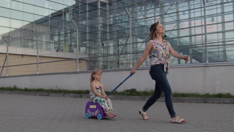 Mother-and-daughter-walking-from-airport-after-vacation.-Woman-rides-baby-child-on-suitcase-luggage