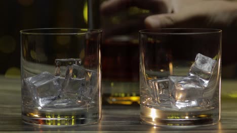 Gießen-Von-Whiskey,-Cognac-Oder-Brandy-Aus-Der-Flasche-In-Gläser-Mit-Eiswürfeln.-Glänzender-Hintergrund